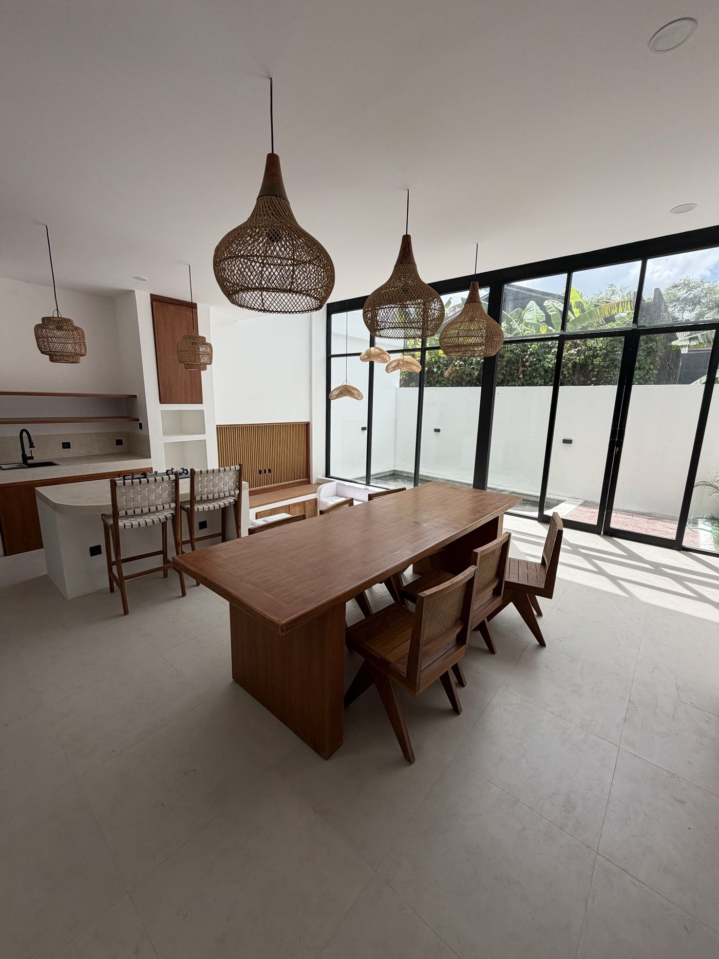 On site, Villa 1: teak dining table and rattan pendants beside full-height black-framed glazing onto the pool courtyard