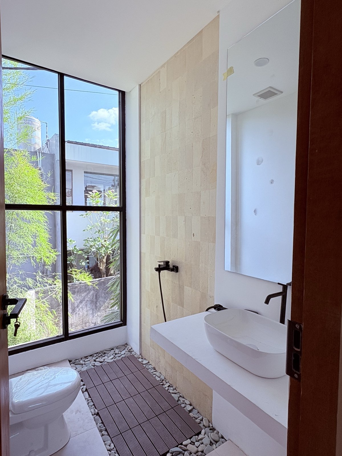 On site: travertine ensuite with vessel basin, black tapware and a timber-and-pebble shower floor beneath full-height black-framed glazing onto the garden